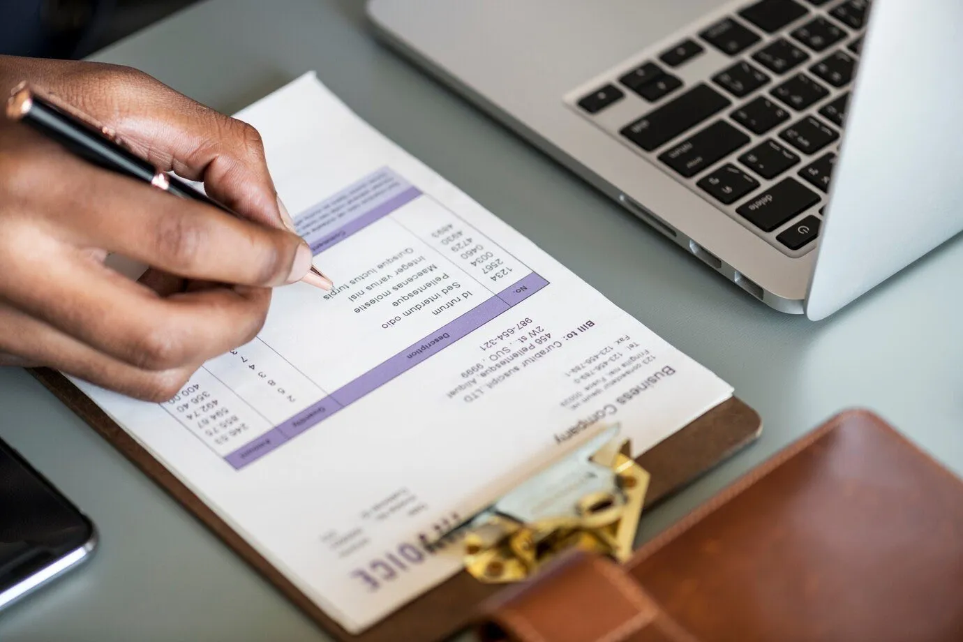 A hand annotating a printed invoice beside a laptop
