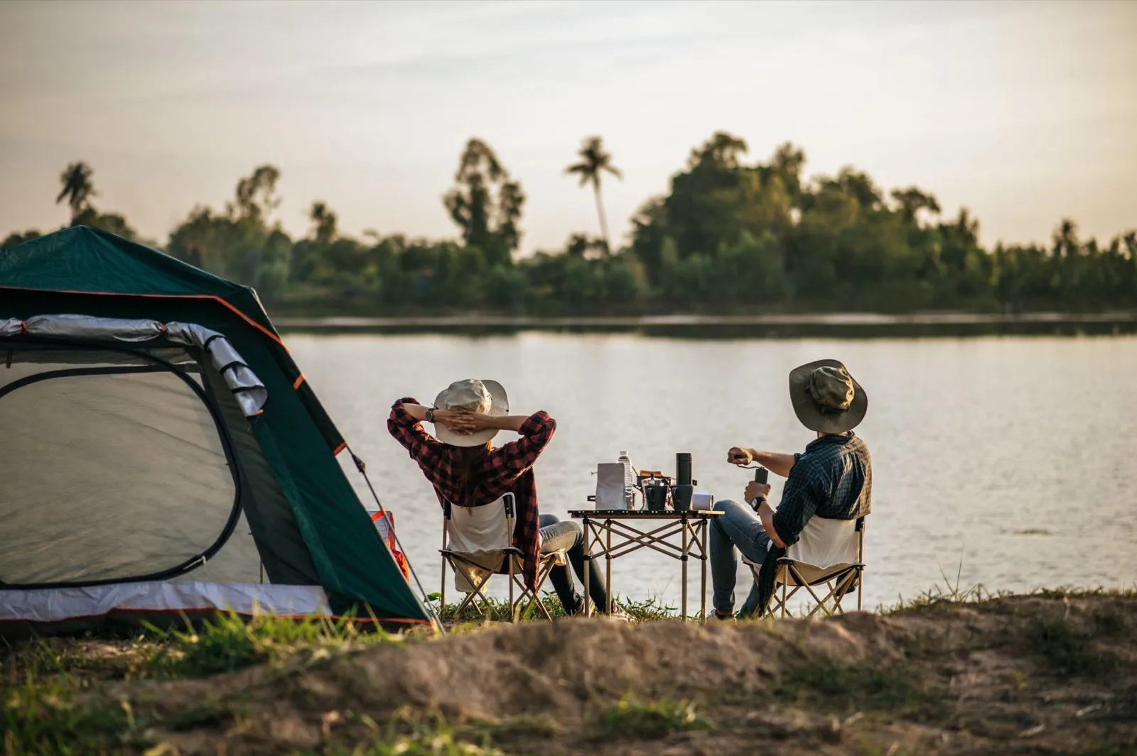 Two people relaxing in camping chairs beside a tent and a lake at sunset